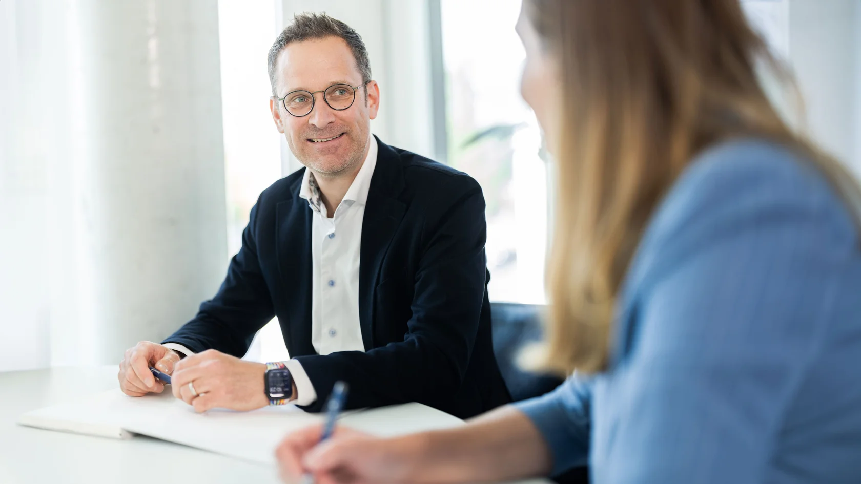 Beratungsgespräch im modernen Büro Lächelnder Mann mit Brille und dunklem Sakko sitzt an einem Tisch und spricht mit einer anderen Person in einem hellen Büro.