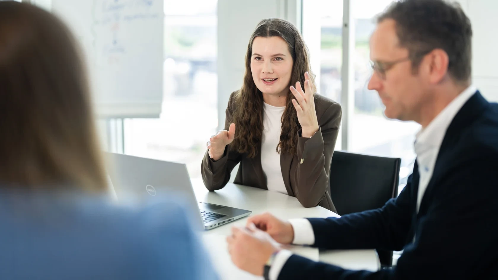 Strategiemeeting mit Fokus auf Erfolg Drei Personen im Gespräch an einem Tisch mit Laptop und Whiteboard im modernen Büro.