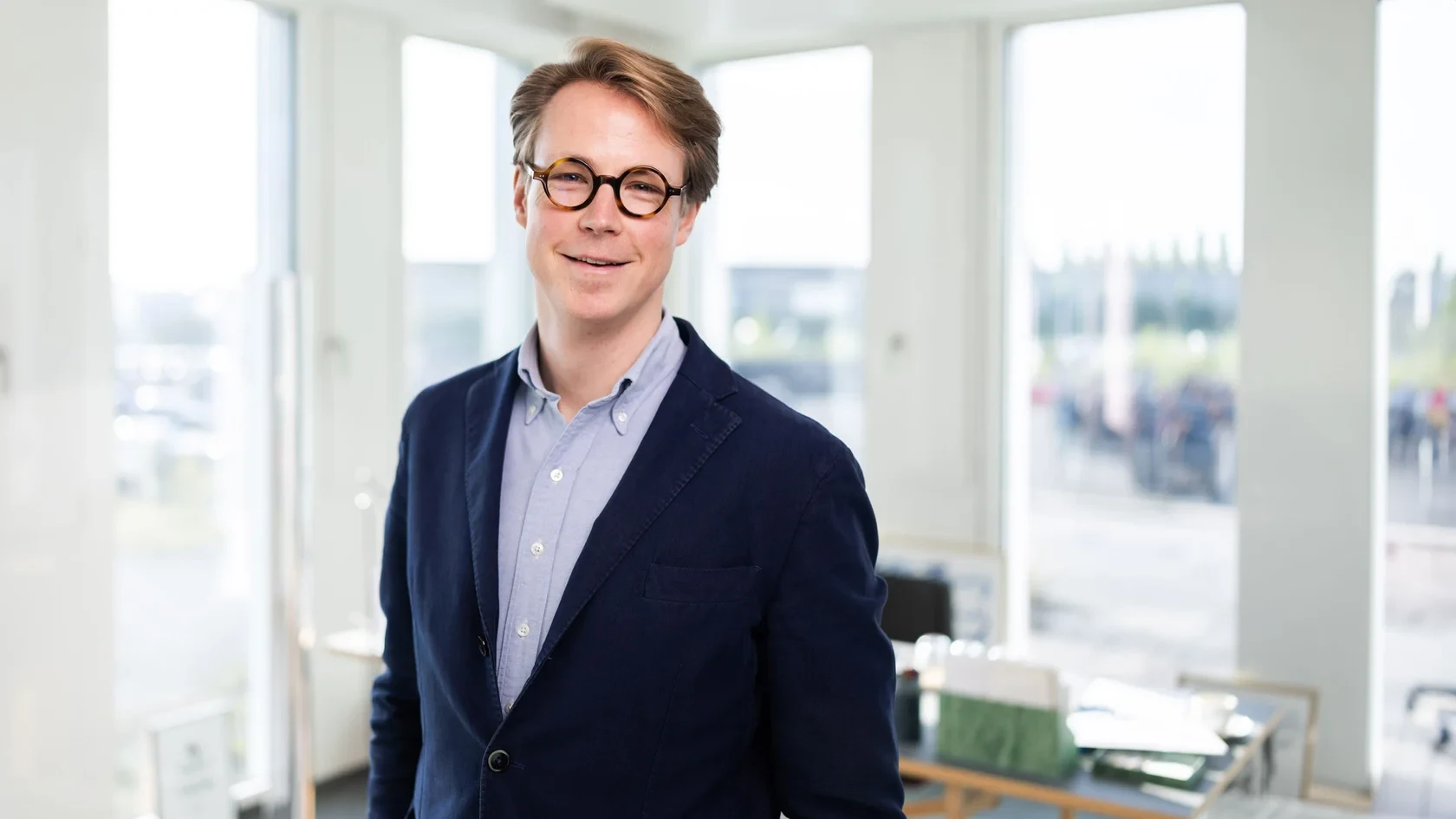 Porträt im lichtdurchfluteten Büro Herr Dr. Sandner mit Brille und Blazer steht in einem hellen Raum mit großen Fenstern im Hintergrund.