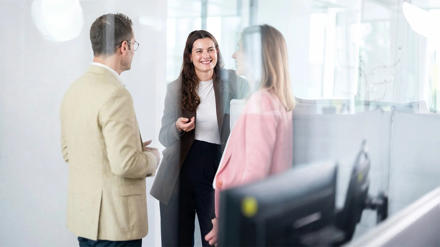 Drei Personen in Business-Kleidung stehen in einem Büro und unterhalten sich. Im Vordergrund ist ein Computermonitor zu sehen.