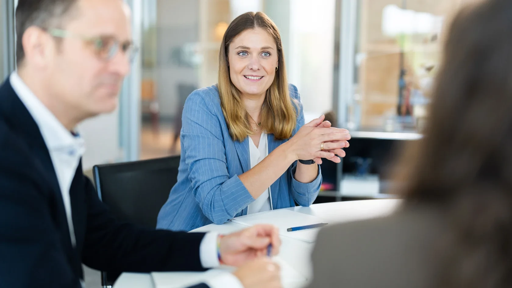 Teammeeting im Büro Eine Gruppe von Personen sitzt um einen Tisch in einem professionellen Bürosetting. Eine Person im blauen Blazer spricht und gestikuliert, während eine andere Person im Vordergrund teilweise sichtbar ist.