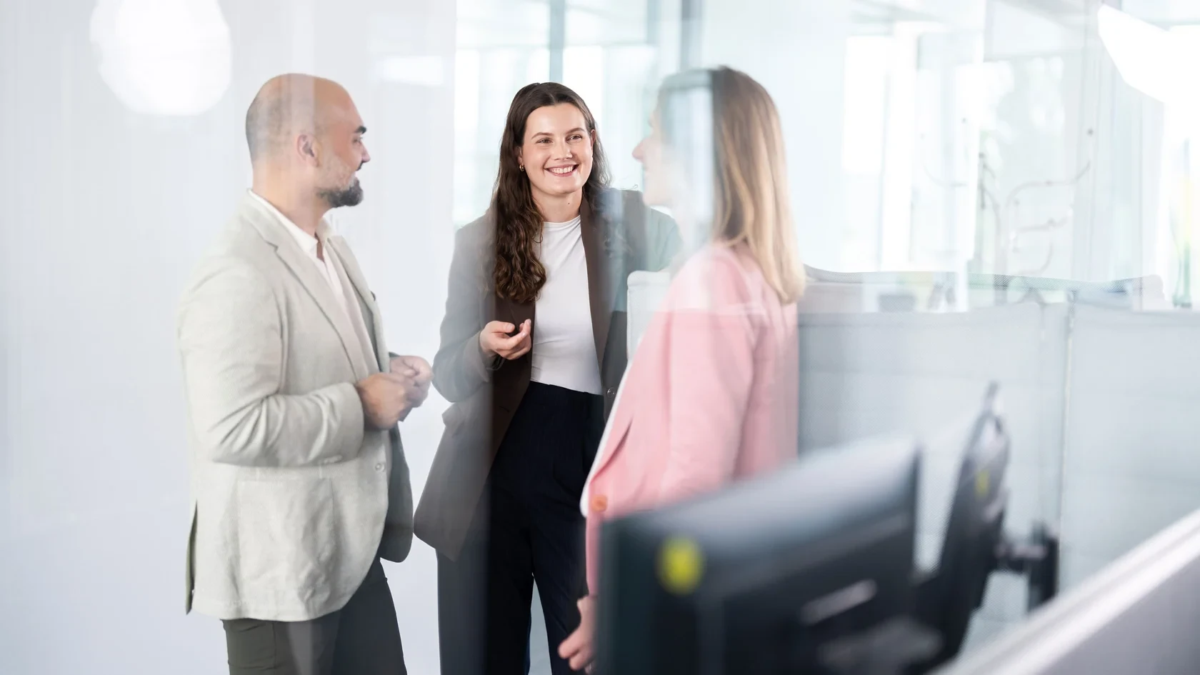 Kollegiale Abstimmung im Arbeitsbereich Drei Personen in Business-Kleidung stehen in einem Büro und unterhalten sich. Im Vordergrund ist ein Computermonitor zu sehen.