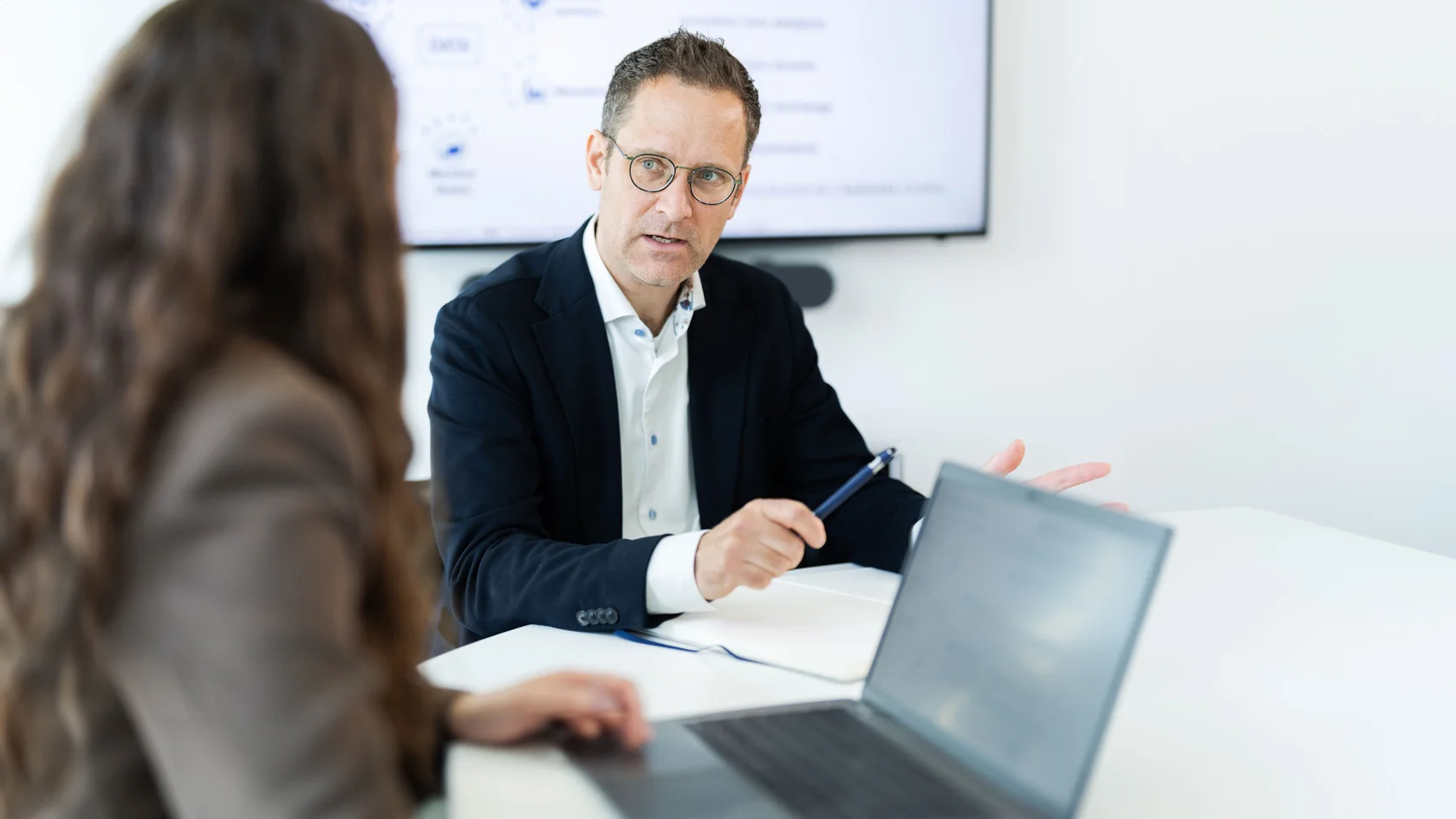 Präsentation im Business-Meeting  Drei Personen in einem Geschäftstreffen, eine Person spricht und hält einen Stift. Im Hintergrund ist ein Präsentationsbildschirm mit Text und Diagrammen zu sehen, auf dem Tisch liegt ein Laptop.