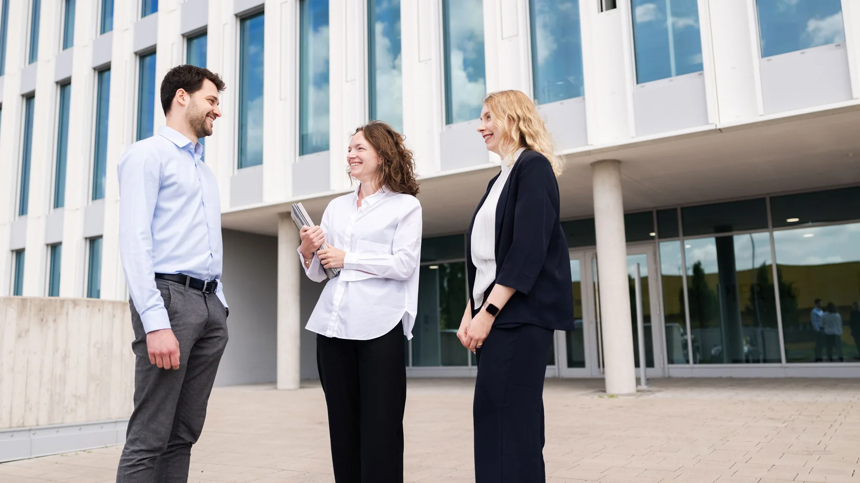 Business Meeting Outside Modern Office Building Drei Personen stehen vor einem modernen Bürogebäude mit großen Fenstern und unterhalten sich.