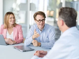 Drei Personen diskutieren an einem Tisch mit Laptops in einem hellen, modernen Büro.