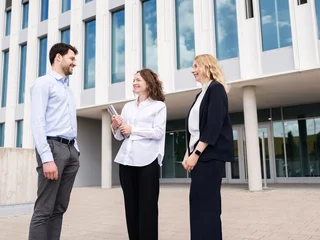 Business Meeting Outside Modern Office Building Drei Personen stehen vor einem modernen Bürogebäude mit großen Fenstern und unterhalten sich.