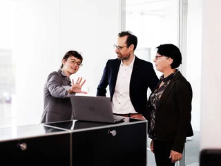 Business Meeting Discussion Drei Personen stehen um einen Laptop auf einem schwarzen Tisch und diskutieren. Eine Person gestikuliert, während die anderen auf den Bildschirm schauen. Im Hintergrund sind Glaswände eines modernen Büros zu sehen.