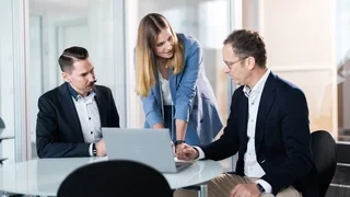 Analyse und Austausch am Laptop Drei Personen in Business-Kleidung sind in einem Büro um einen Tisch versammelt. Eine Person steht und zeigt auf den Laptop-Bildschirm, während die anderen beiden sitzen und aufmerksam zuschauen.