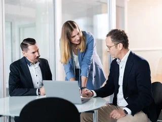Analysis and discussion at the laptop. Three people in business attire are gathered around a table in an office. One person is standing and pointing at the laptop screen, while the other two are seated and watching attentively.