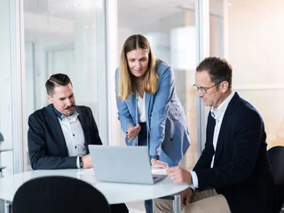 Strategieplanung am runden Tisch Drei Personen in Business-Kleidung stehen und sitzen um einen runden Tisch mit einem Laptop in einem modernen Büro. Eine Person steht und zeigt auf den Bildschirm, während die anderen beiden aufmerksam zusehen.