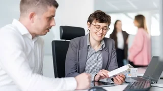 Zwei Mitarbeitende im Büro im Gespräch; eine Frau zeigt einem Kollegen eine Medikamentenverpackung, während im Hintergrund zwei weitere Personen stehen und sich unterhalten.