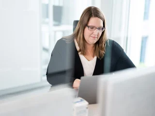 Konzentriertes Arbeiten im modernen Büro Eine Person sitzt an einem Schreibtisch in einem modernen Büro und arbeitet an einem Laptop. Das Gesicht ist unscharf, im Hintergrund sind große Fenster mit Tageslicht zu sehen.