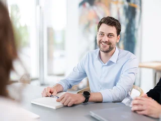 Notizen im Meeting Eine Person in einem hellblauen Hemd sitzt an einem Tisch mit einem offenen Notizbuch, hält einen Stift in der rechten Hand und trägt eine schwarze Armbanduhr. Im Hintergrund sind weitere Personen unscharf zu erkennen.