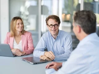 Business-Meeting am Fensterplatz Drei Personen bei einem Meeting mit Laptops in einem hellen Büro mit großen Fenstern.