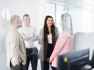 Kollegiale Abstimmung im Arbeitsbereich Drei Personen in Business-Kleidung stehen in einem Büro und unterhalten sich. Im Vordergrund ist ein Computermonitor zu sehen.
