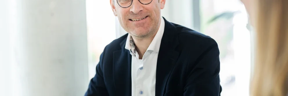 Lächelnder Mann mit Brille und dunklem Sakko sitzt an einem Tisch und spricht mit einer anderen Person in einem hellen Büro.