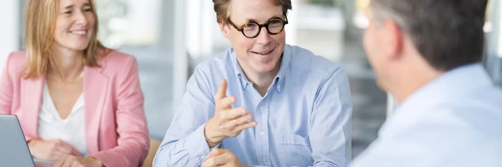 Drei Personen diskutieren an einem Tisch mit Laptops in einem hellen, modernen Büro. Drei Personen diskutieren an einem Tisch mit Laptops in einem hellen, modernen Büro.