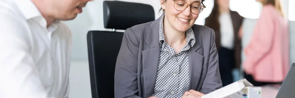 Fokusgespräch im Büro mit Hintergrundaustausch Zwei Personen sitzen an einem Schreibtisch in einem professionellen Umfeld, eine davon hält ein Dokument. Im Hintergrund unterhalten sich zwei weitere Personen. Auf dem Tisch liegen Laptops und Büromaterialien.