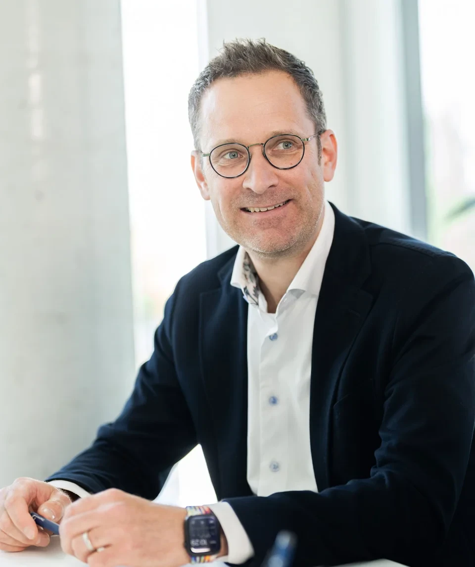 Lächelnder Mann mit Brille und dunklem Sakko sitzt an einem Tisch und spricht mit einer anderen Person in einem hellen Büro.