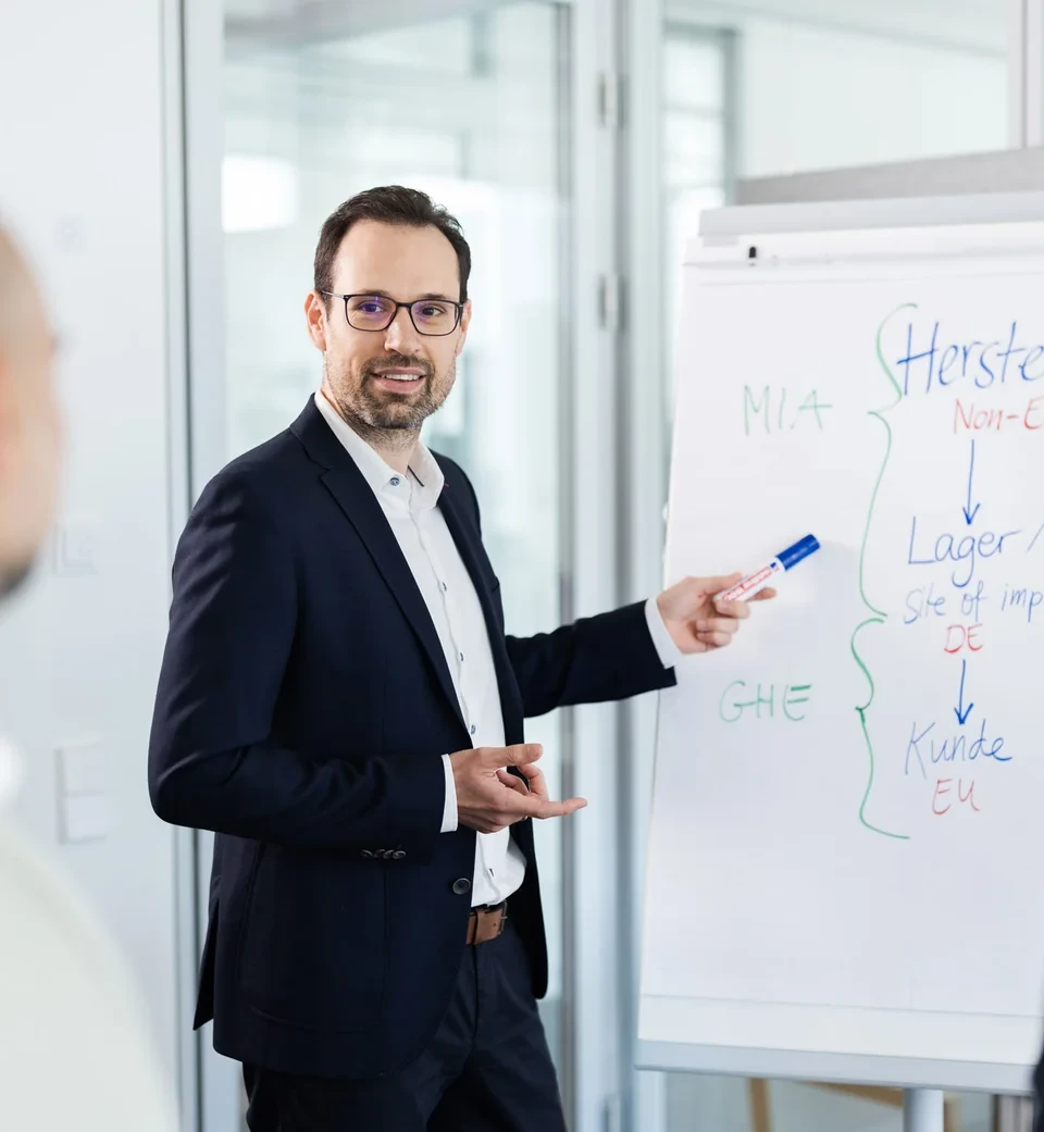 Ein Mann im Anzug hält eine Präsentation für zwei weitere Personen und nutzt dabei ein Flipchart mit Stichpunkten zu Import- und Lagerprozessen.