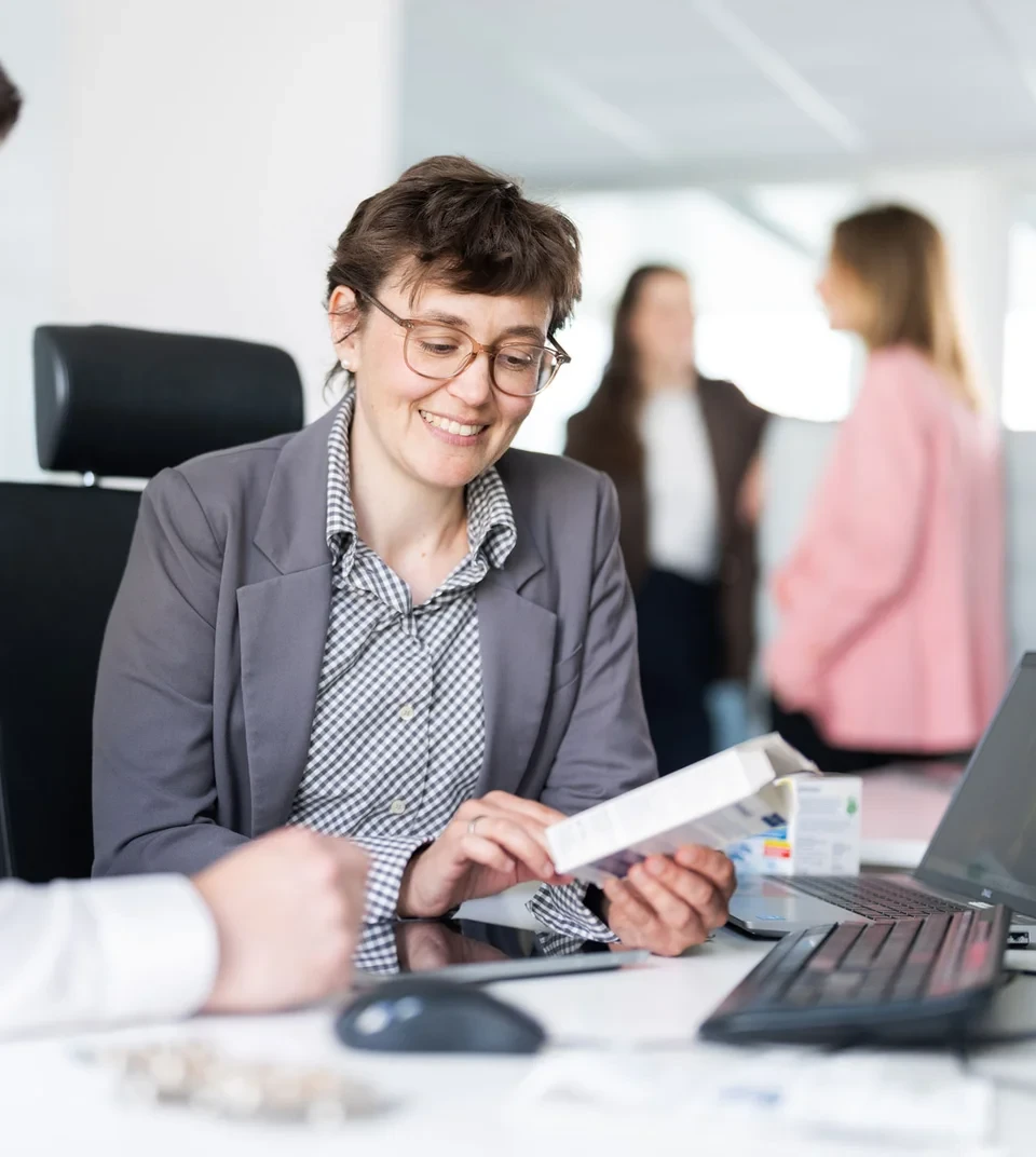 Zwei Personen sitzen an einem Schreibtisch in einem professionellen Umfeld, eine davon hält ein Dokument. Im Hintergrund unterhalten sich zwei weitere Personen. Auf dem Tisch liegen Laptops und Büromaterialien.